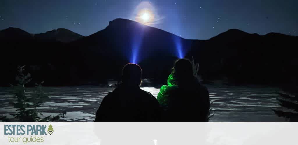 Two people gaze at a serene night sky by a lake, silhouetted against the backdrop of mountains. A bright moon shines above, casting reflections on the water. Stars dot the sky, and beams of light point towards the moon, creating a peaceful and contemplative scene. Dense foliage frames the image, capturing the essence of a tranquil outdoor adventure.