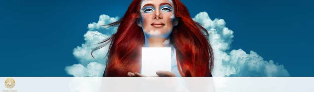 A person with long red hair stands against a vivid blue sky and fluffy white clouds. They hold a glowing rectangular object in both hands, emitting a bright light on their face. The person's makeup is artistic and bold, enhancing the mystical atmosphere of the scene.