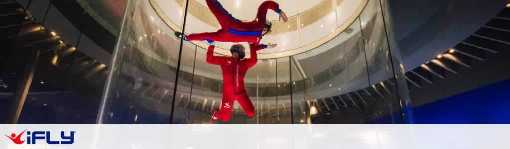 Two people in red flight suits experience indoor skydiving inside a wind tunnel at iFLY. One person is elevated above the other, both wearing helmets for safety. The tunnel is enclosed with clear walls, and the interior is well-lit, highlighting the dynamic movement. The environment gives a sense of thrilling flight and excitement.