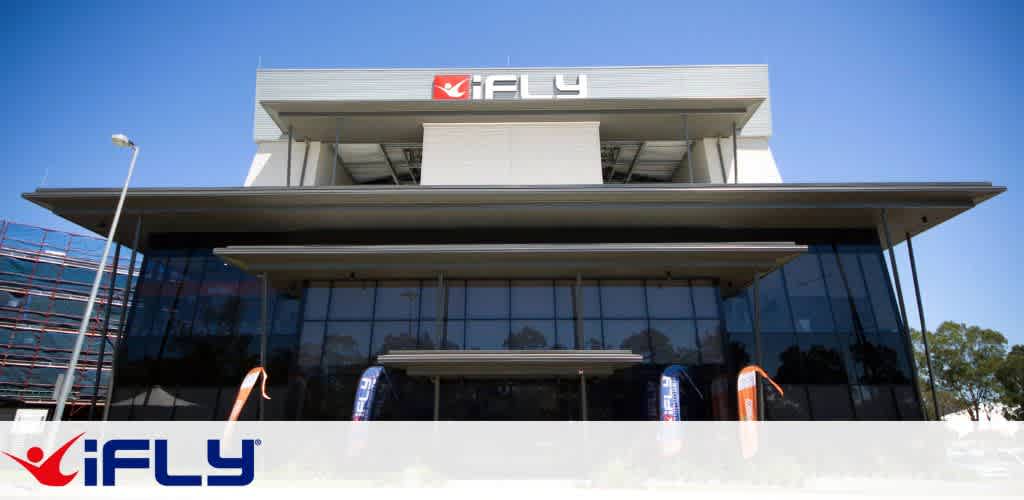 The image shows a modern iFLY building under a clear blue sky. The structure features large glass windows and a prominent gray facade, with the iFLY logo displayed at the top. Two colorful banners flank the entrance, adding a vibrant touch to the sleek, contemporary design. The setting suggests an inviting and dynamic venue, possibly for indoor skydiving experiences, aimed at drawing in thrill-seekers and adventure enthusiasts.