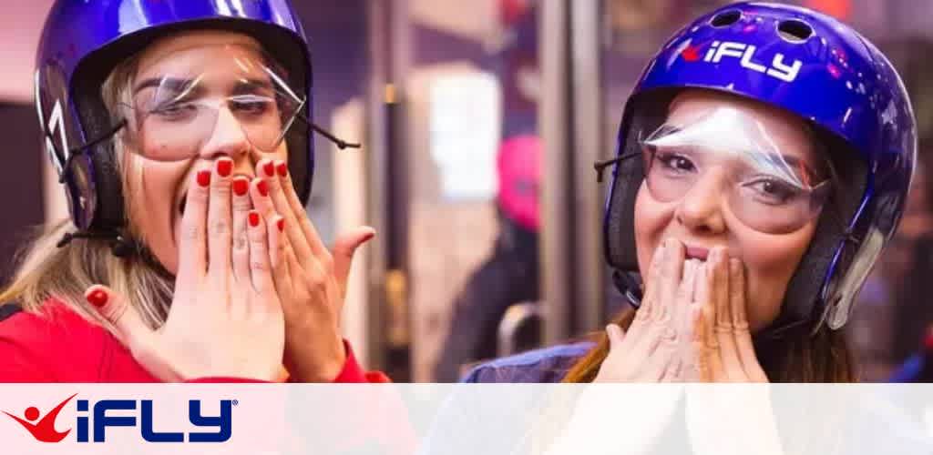 Two people wearing blue helmets and protective goggles are standing indoors. Both have their mouths partially covered by their hands, appearing excited or surprised. The iFLY logo is visible on their helmets and at the bottom of the image, suggesting an indoor skydiving experience. The setting has a vibrant and energetic atmosphere.