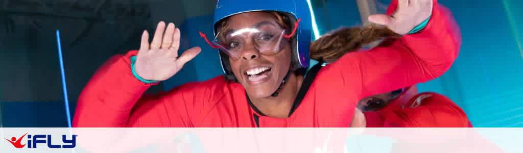 A person wearing a red flight suit and blue helmet is floating in a vertical wind tunnel at a skydiving facility. They have a joyful expression, with arms extended forward. The background is a blurred interior with blue lights. A logo for iFLY is visible in the bottom left corner.