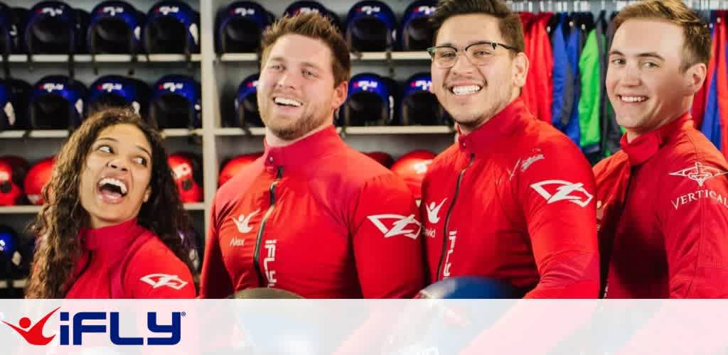 A group of four people in red flight suits are smiling and standing together, ready for an indoor skydiving experience at iFly. Shelves with helmets and colorful suits are visible in the background, adding to the lively atmosphere. The iFly logo is at the bottom, reflecting the excitement and camaraderie of the activity.
