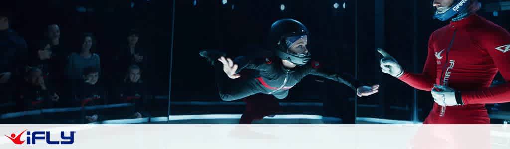 Image shows an indoor skydiving experience at iFLY. A person in a black flight suit and helmet is hovering mid-air inside a vertical wind tunnel. An instructor in a red suit guides them with hand signals. In the background, spectators watch from behind a glass barrier. The setting is well-lit with a focus on the flyer and instructor.