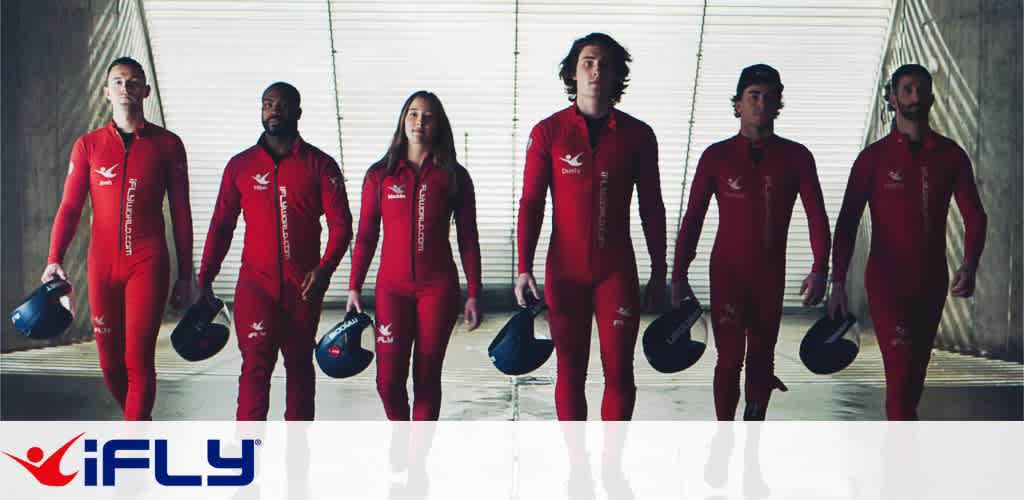A team of six individuals in red jumpsuits walks confidently towards the camera. They carry helmets and stand in a sunlit indoor space with metal walls and large windows, suggesting a skydiving or wind tunnel environment. The iFLY logo is in the bottom corner, indicating a connection to indoor skydiving activities.