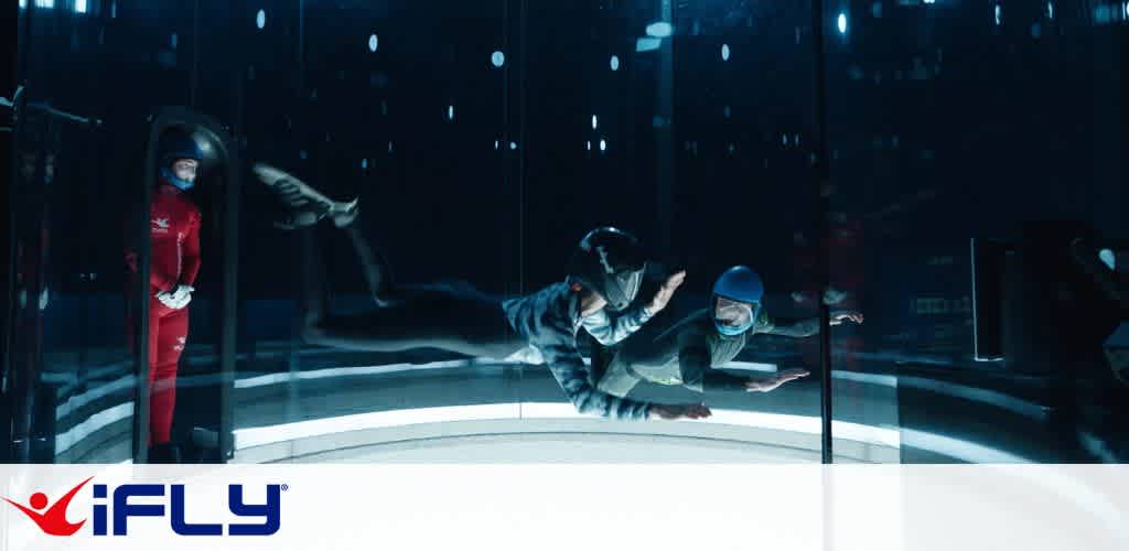 Individuals experience indoor skydiving in a vertical wind tunnel at iFLY. Two people are suspended in mid-air, practicing maneuvers and guided by an instructor in a red suit standing nearby. All are wearing helmets and safety gear. The setting is dimly lit, highlighting the excitement and thrill of flight simulation. A logo is visible at the bottom, emphasizing the brand.