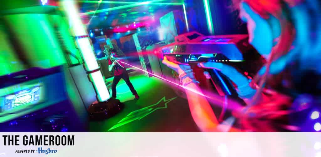 Colorful laser tag arena featuring players engaged in a game. Bright neon lights in blue, green, and pink glow throughout the space, creating an exciting and dynamic atmosphere. Participants aim laser guns amid glowing obstacles, enhancing the futuristic vibe. Text at the bottom reads The Gameroom powered by Hasbro.