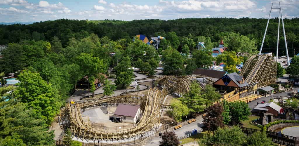 Aerial view of a lush, green amusement park featuring a wooden roller coaster winding through the trees. Surrounding attractions include colorful structures and rides, creating a vibrant atmosphere. The sky is blue with scattered clouds, adding to the lively and inviting scene.