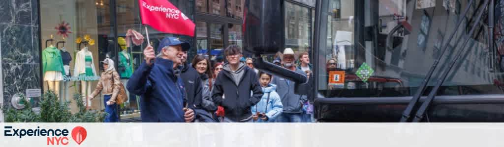 A tour guide holds a red flag labeled Experience NYC as a group of people stands beside a tour bus on a city street. Behind them, a store window displays colorful clothing. The participants appear to be diverse, dressed in various jackets suitable for cool weather. The bus is black, and the area is bustling with city life.