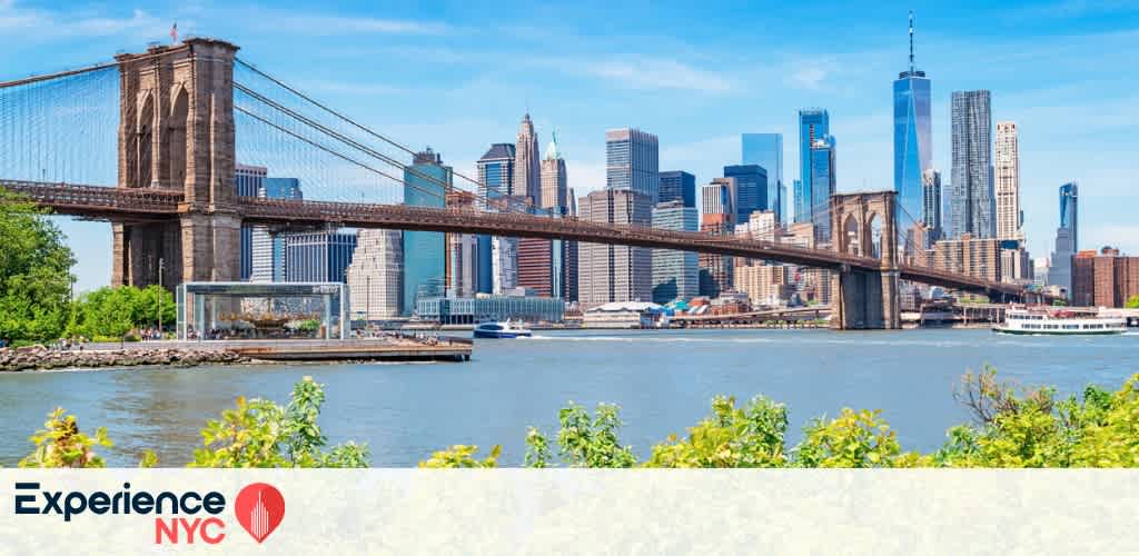 View of the Brooklyn Bridge spanning the East River with the iconic Manhattan skyline in the background. Tall skyscrapers rise against a clear blue sky. In the foreground, lush green plants frame the scene. Experience NYC text with a red location pin icon is at the bottom left.