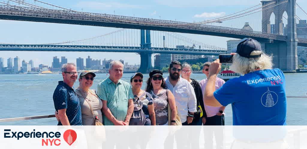 A group of people poses for a photo with the Brooklyn Bridge and New York City skyline in the background. The scene is sunny, and they are on a waterfront walkway. A person stands to the right, using a smartphone to capture the moment. This image promotes Experience NYC with a logo in the bottom left corner.