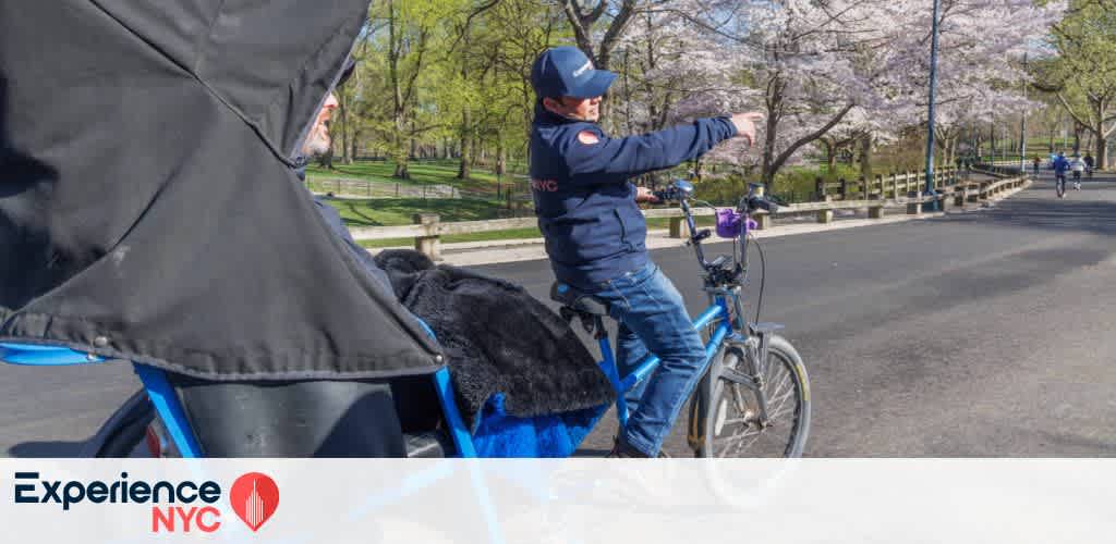 A tour guide on a pedicab points out a scenic view in a park lined with blossoming cherry trees. The road is clear, with another cyclist in the distance. The setting is calm and picturesque, perfect for exploring on a sunny day. A logo for Experience NYC is displayed in the corner.