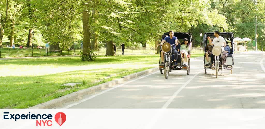 A sunny day in a park with lush green trees lining a paved path. Two pedicabs, each carrying passengers, are ridden side by side down the road. People are enjoying a leisurely ride, surrounded by vibrant greenery. In the foreground, the Experience NYC logo is displayed at the bottom left corner.