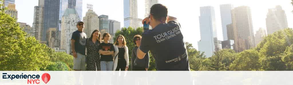 A group of people is being photographed in front of a lush green park with a city skyline in the background. The photographer wears a tour guide shirt. Sunlight softly illuminates the tall buildings and the group, creating a bright and inviting atmosphere. The park and cityscape suggest a vibrant urban experience, emphasizing the blend of nature and city life. The image conveys an essence of exploration and discovery.
