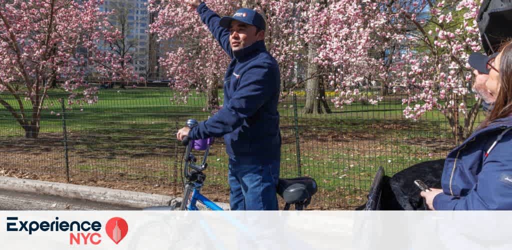 A person points towards the sky while standing beside a bicycle in a park. They are surrounded by trees blooming with pink flowers under a clear blue sky. Another person, partially visible, listens to them. The scene exudes a sense of exploration and enjoyment of a sunny day amidst beautiful nature. The logo 'Experience NYC' is displayed in the lower left corner.