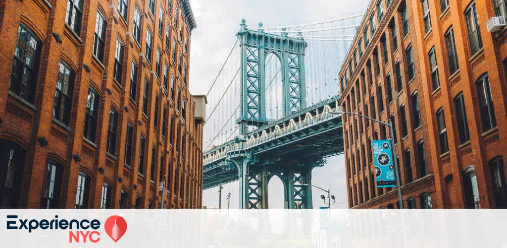 Image of the Manhattan Bridge framed by iconic brick buildings in New York City. The structure stands prominently against the sky, showcasing its architectural beauty. The scene reflects the vibrant urban atmosphere of NYC. The Experience NYC banner is visibly placed in the bottom left corner.