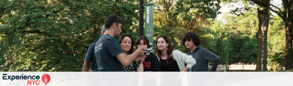 A group of five people stand together in a lush, green park setting, with trees and sunlight filtering through. The person in the front holds a phone, showing something to the others who seem engaged and smiling. A signpost and a pathway are visible in the background, enhancing the vibrant outdoor atmosphere. The image conveys a sense of camaraderie and enjoyment in a natural urban environment. The Experience NYC logo is in the lower left corner.
