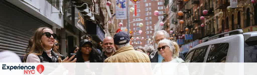 A group of people is standing on a bustling city street, surrounded by tall buildings and colorful hanging lanterns. They appear to be in a lively discussion or tour, with one person gesturing as they speak. The scene captures the vibrant energy and diversity of a city experience. In the foreground, two people wear sunglasses. Hanging lights add to the festive atmosphere.