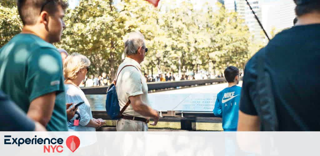A group of people gather around an informative display in an outdoor setting, surrounded by trees. Various individuals of different ages are engaged, some reading the display, while others observe their surroundings. The scene is bright and sunny, suggesting a warm day. The image promotes the Experience NYC brand, visible in the corner with a red location pin design.