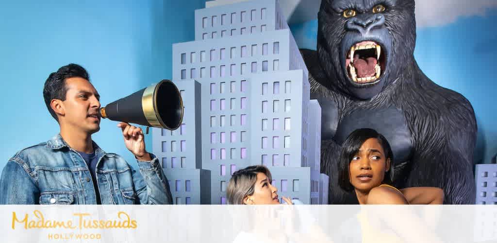 A person with a megaphone standing next to two women and a large gorilla sculpture in front of stylized city buildings inside Madame Tussauds Hollywood.