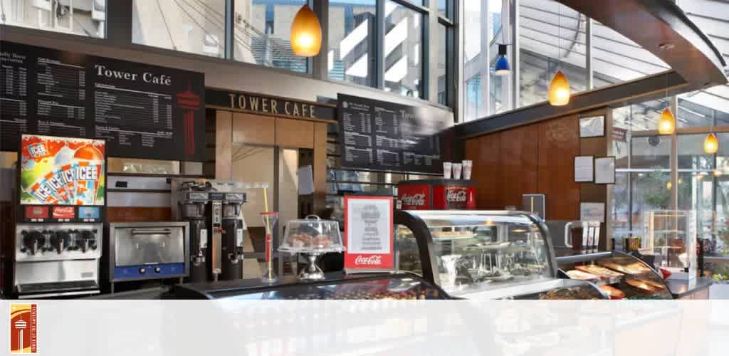 The image shows the interior of a modern café with a large menu board displaying options. A counter at the front features a glass display filled with food items and drink machines, including an ICEE machine. Warm lighting fixtures hang from the ceiling, and large windows in the background allow natural light to fill the space. The ambiance is inviting and contemporary.