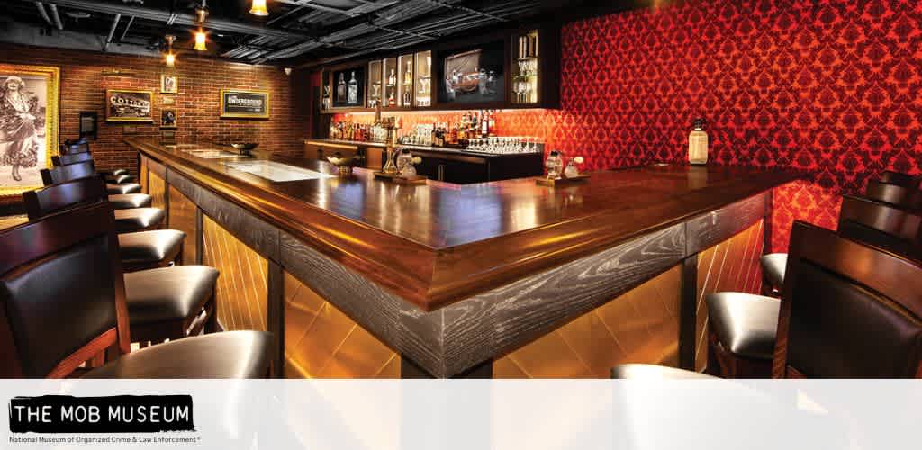 The image shows an upscale bar with a wooden counter and leather bar stools. The decor features a red patterned wall and framed vintage photos. The lighting is warm, creating an inviting atmosphere. Bottles and glasses are neatly arranged behind the bar. The setting appears to be in a stylish, retro-themed space.