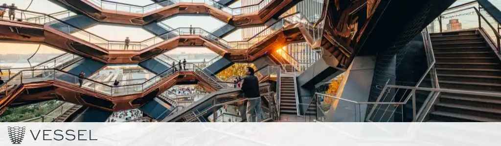 A view of the Vessel, a striking architectural structure featuring interwoven staircases and platforms. People explore different levels, surrounded by sunset light reflecting off the metallic surfaces. The design offers unique perspectives and overlooks a cityscape backdrop.