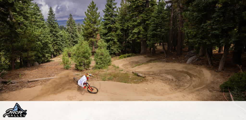 Mountain biker navigates a winding dirt trail surrounded by lush green trees under a cloudy sky at Snow Valley. The path curves through the dense forest, providing an adrenaline-fueled descent. Located in a picturesque natural setting, this trail offers a thrilling ride for outdoor enthusiasts. The scene captures the excitement and beauty of mountain biking amidst towering pines and a rugged landscape.