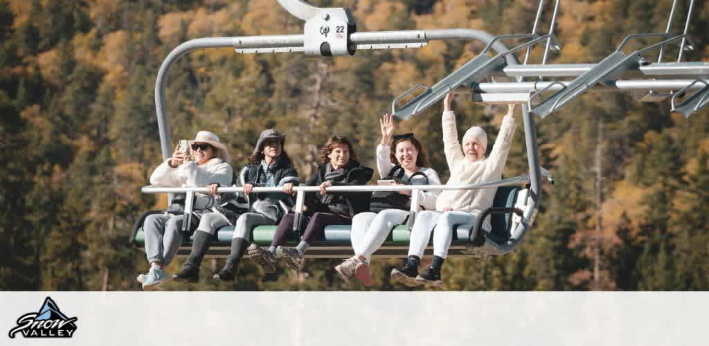 A group of five people enjoy a scenic ride on a ski lift at Snow Valley. They sit comfortably bundled in warm clothes, and some wave cheerfully while others capture the moment with their phones. The background shows a lush forest with autumn leaves in warm tones. The relaxed atmosphere highlights a fun day in nature.