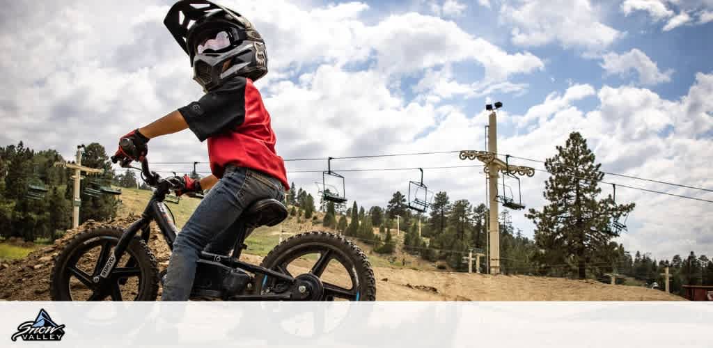 A young person is riding a BMX bike on a dirt trail, wearing a helmet and gloves. The background features a ski lift and a forested area under a partly cloudy sky. The scene captures an adventurous outdoor activity in a mountainous location, indicating a sense of excitement and exploration. Snow Valley logo is visible in the corner.