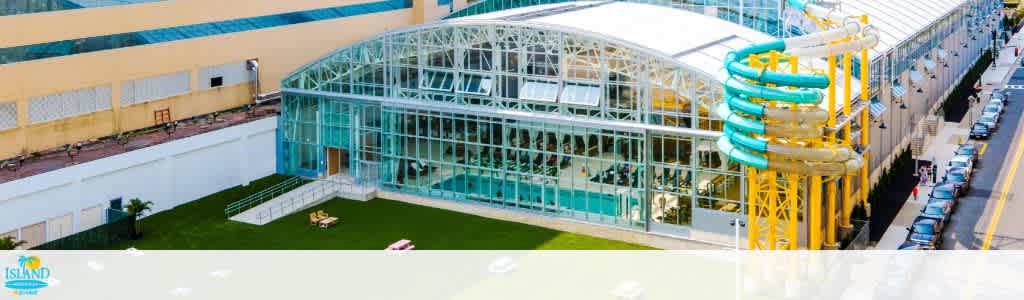 The image shows a large glass-roofed building housing a pool, surrounded by a grassy area with lounge chairs. A brightly colored spiral water slide extends from the roof down the side. The structure is adjacent to a street with parked cars, set against a backdrop of yellow and white buildings. The design suggests a recreational or leisure facility.