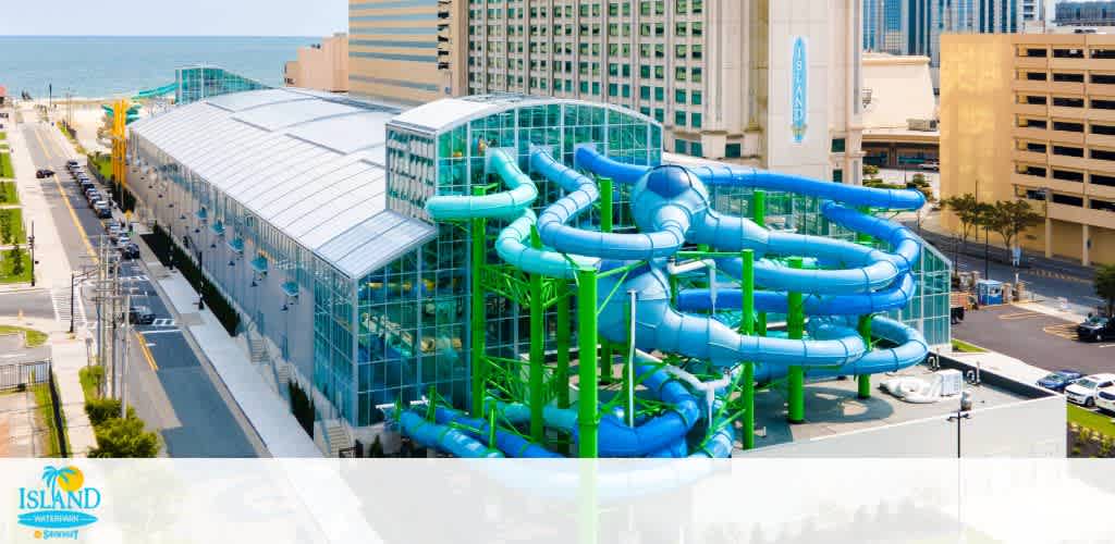 A vibrant water park surrounded by city buildings with multiple intertwined blue and green water slides visible. The indoor section has a large glass roof, and the park is located near the ocean. The park offers a mix of thrilling water attractions and urban views, providing a fun getaway in a coastal city setting.