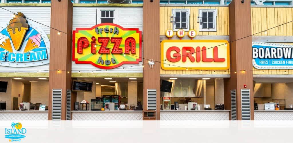 Exterior view of a vibrant food court at Island Waterpark. It features colorful signage for ice cream, fresh hot pizza, a grill, and boardwalk-style food offerings such as fries and chicken fingers. Bright lights and warm colors create a welcoming atmosphere. Perfect for quick meals and treats while enjoying the waterpark.