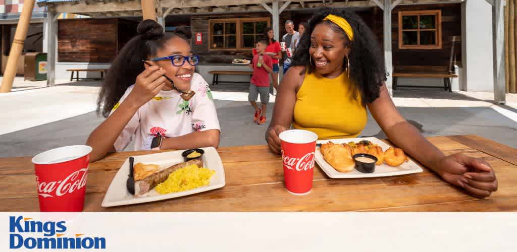Two people sitting at a table with meals and Coca-Cola cups, smiling at each other in an outdoor amusement park setting.