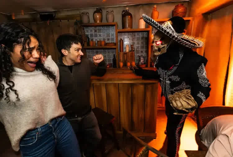 Two people react in fear and surprise inside a dimly lit room decorated with rustic pottery and wooden furniture, as a person dressed in a skeleton costume with glowing red eyes and a sombrero approaches them menacingly from behind a bar counter. The scene appears to be part of a themed haunted attraction or escape room experience.