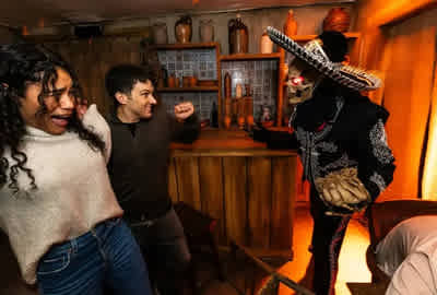 Two people react in fear and surprise inside a dimly lit room decorated with rustic pottery and wooden furniture, as a person dressed in a skeleton costume with glowing red eyes and a sombrero approaches them menacingly from behind a bar counter. The scene appears to be part of a themed haunted attraction or escape room experience.