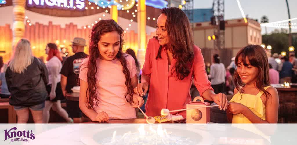 Three people roasting marshmallows around a fire pit at an amusement park in the evening.