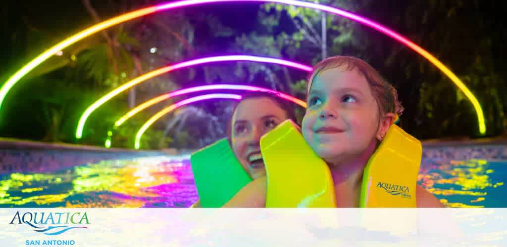 A child and adult in a pool at night with colorful light arcs in the background.