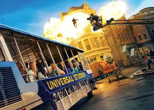 A Universal Studios tour bus passes through a lively movie set. Visitors watch as a thrilling scene unfolds with a stuntman launched into the air amidst a fiery explosion. Film equipment and crew are visible, showcasing the excitement of behind-the-scenes action.