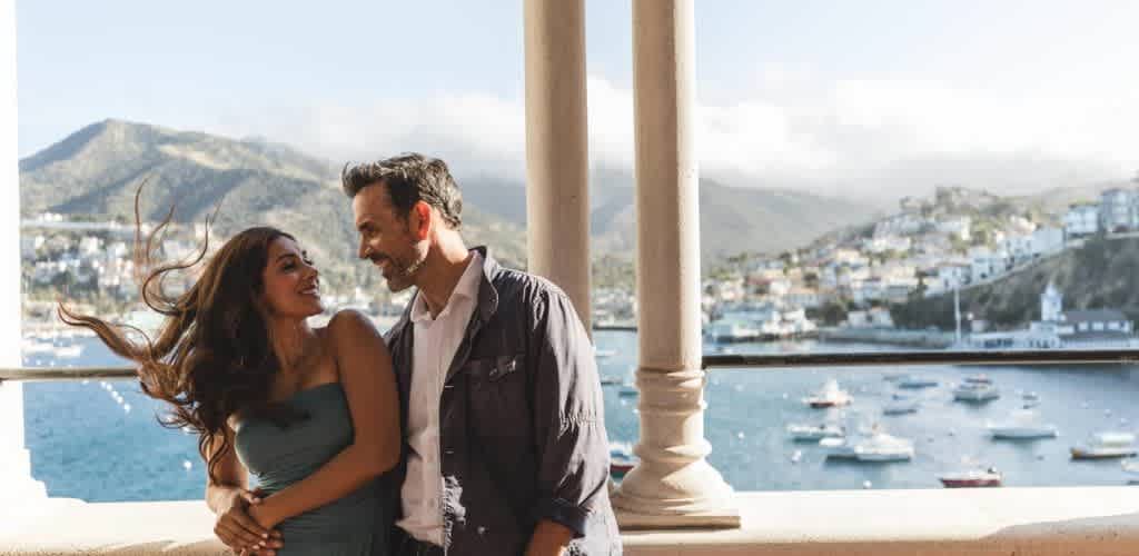 A smiling couple stands on a balcony overlooking a scenic bay with boats floating on the water. Hills with houses are visible in the background. The woman wears a strapless dress, and the man is in a casual shirt and jacket. The atmosphere is sunny and relaxed, suggesting a joyful and carefree moment.
