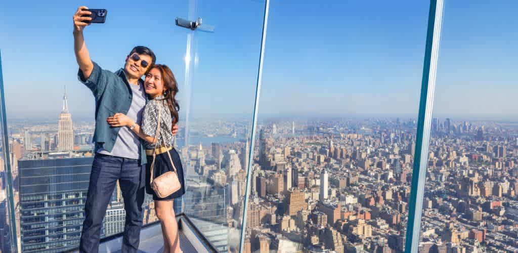A couple smiles while taking a selfie at a high observation deck with glass railings, offering a panoramic view of a sprawling cityscape. The skyline features iconic skyscrapers under a clear blue sky, creating a vibrant backdrop for their shared moment.