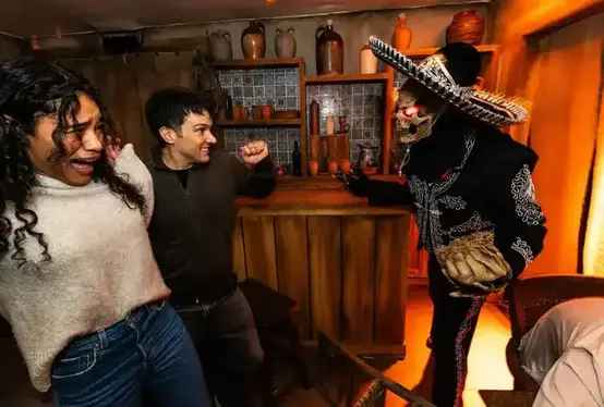 Two people react in fear and surprise inside a dimly lit room decorated with rustic pottery and wooden furniture, as a person dressed in a skeleton costume with glowing red eyes and a sombrero approaches them menacingly from behind a bar counter. The scene appears to be part of a themed haunted attraction or escape room experience.