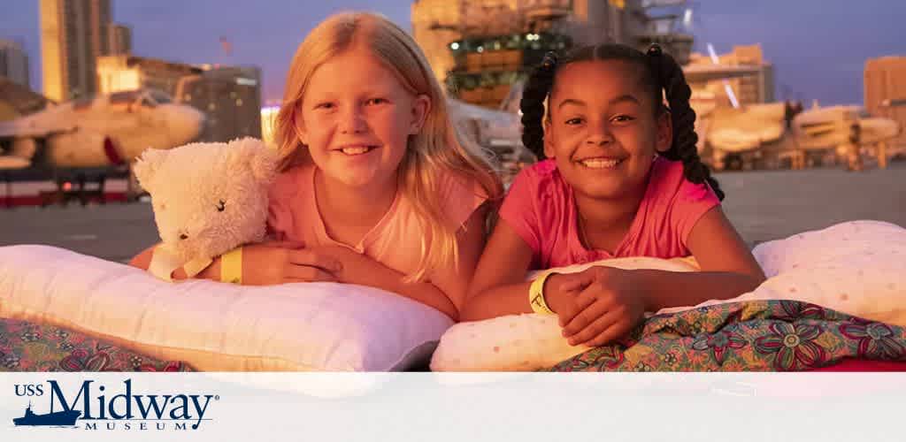 Two girls lying on pillows on the deck of the USS Midway Museum, in front of aircraft and cityscape.