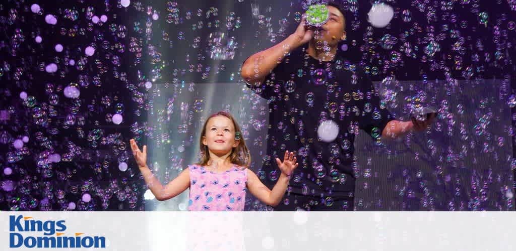 A young girl and a person with a microphone stand among bubbles on stage, under bright lights.