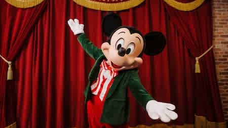 Mickey Mouse wearing a green holiday jacket and red-striped shirt, posing cheerfully in front of a red velvet stage curtain at Disneyland.