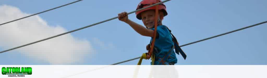 A child wearing a red helmet and blue shirt is ziplining on a clear day. They are securely harnessed and smiling, holding onto the cable with one hand. The sky is bright blue with scattered clouds. Gatorland Orlando Florida is text on bottom left.
