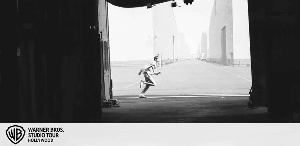 A person runs between large buildings on a studio backlot in black and white.