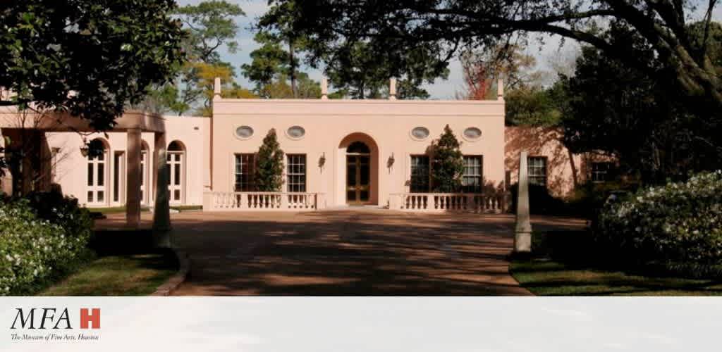 Elegant pale pink building with arched entrance and symmetrical windows sits amid lush greenery. Tall trees provide shade, creating a serene atmosphere. The entrance is framed by well-manicured bushes and flanked by two pillars. The foreground features a driveway, leading towards the entrance. A logo at the bottom reads MFAH, Museum of Fine Arts, Houston.