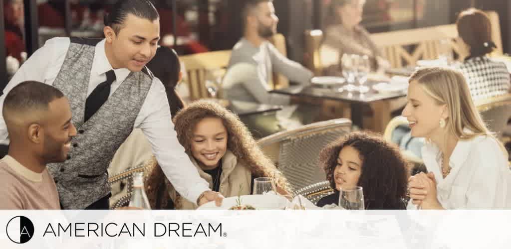 A server in a gray vest serves a smiling family at a restaurant table.