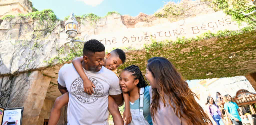 A family of five smiling and enjoying time together outdoors in front of a themed attraction with a sign that says the adventure begins